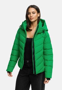 Femme portant une veste rembourrée vert vif avec capuche, un haut noir et un pantalon noir, se tenant devant un fond gris clair uni.