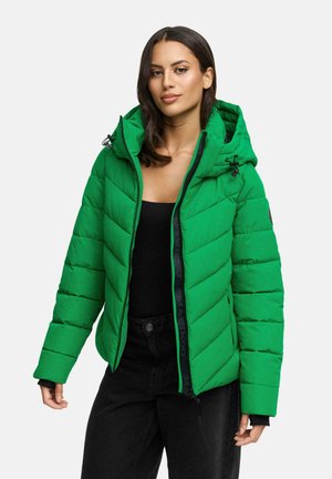 Femme portant une veste rembourrée vert vif avec capuche, un haut noir et un pantalon noir, se tenant devant un fond gris clair uni.