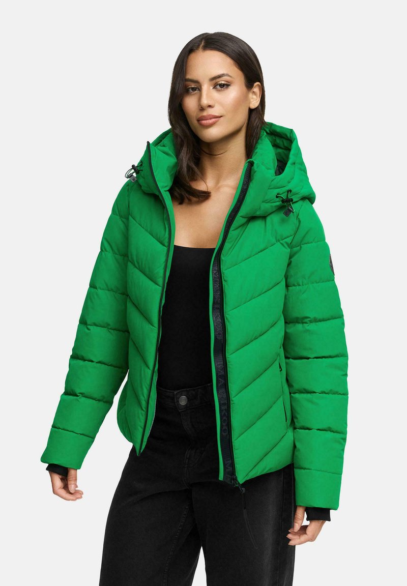 Femme portant une veste rembourrée vert vif avec capuche, un haut noir et un pantalon noir, se tenant devant un fond gris clair uni.