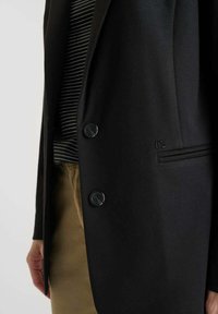 Blazer noir au toucher lisse, doté de deux boutons noirs, d'une doublure à rayures discrètes et d'une poche latérale, porté sur un pantalon kaki.