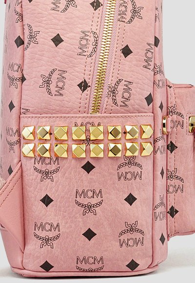 Materiale texturizzato rosa con loghi MCM neri, zip dorata e dettagli in accessori borchiati sul pannello frontale. Presenta elementi di design geometrico.