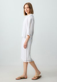 Weißes Leinenoutfit mit einem kurzärmeligen Oberteil mit braunen Streifen und einem knielangen Rock. Kombiniert mit braunen Flip-Flops. Minimalistisches Design.