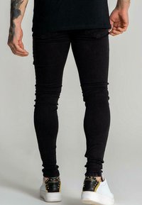 Svarta skinny jeans i texturerat tyg, med en figurnära design och fransdetalj vid midjan, matchade med vita och svarta sneakers.