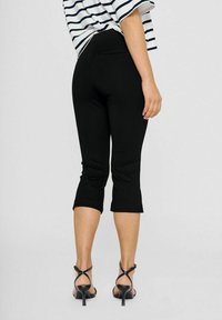 Zwarte cropped broek met een getailleerd ontwerp, voorzien van een gladde textuur en een lengte tot halverwege de kuit. Gecombineerd met zwarte hoge hakken.
