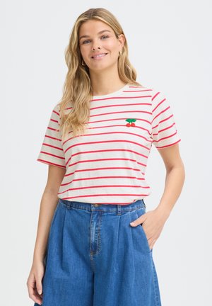Femme blonde souriante, portant un t-shirt blanc à manches courtes avec des rayures horizontales rouges et une broderie de cerises, associée à un jean bleu taille haute.