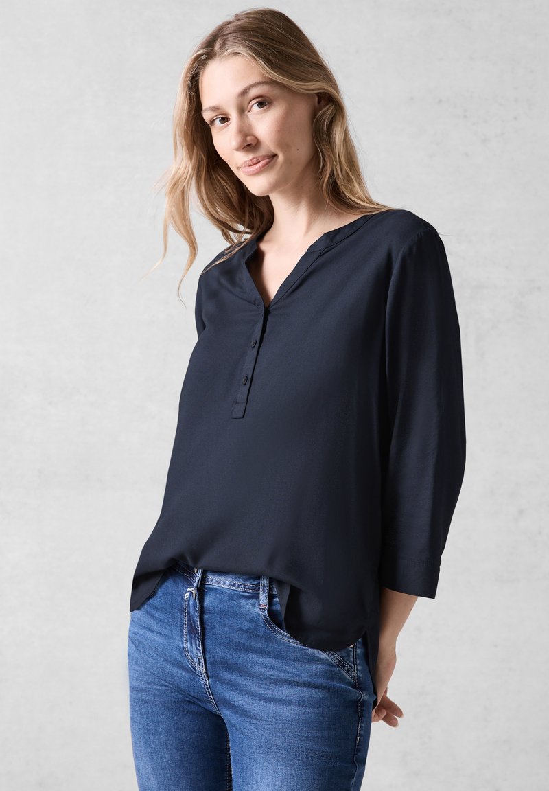 Donkerblauwe, lange mouwen blouse met een V-hals en drie knopen aan de voorzijde, gemaakt van zachte stof, gecombineerd met blauwe jeans.
