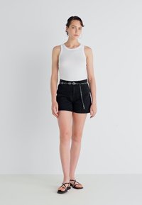 Un maieu alb asortat cu pantaloni scurți negri, evidențiat de un brâu decorativ și o lanț argintiu. Modelul poartă sandale negre cu bretele. Fundal simplu.