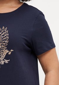 Tumši zila kokvilnas t-krekls ar detalizētu zelta dizainu un dārgakmeņiem priekšpusē; īsas piedurknes un klasiskā apaļā kakla izgriezums.
