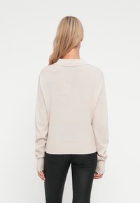 Pull en tricot beige avec un col et un ourlet côtelé, doté de manches longues et d'une coupe décontractée, porté avec un pantalon noir.