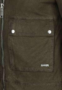 Perforeerde groene jas met een rits aan de voorkant, zilverkleurige drukknopen op de zakken en een klein logo-label aan de onderkant links.