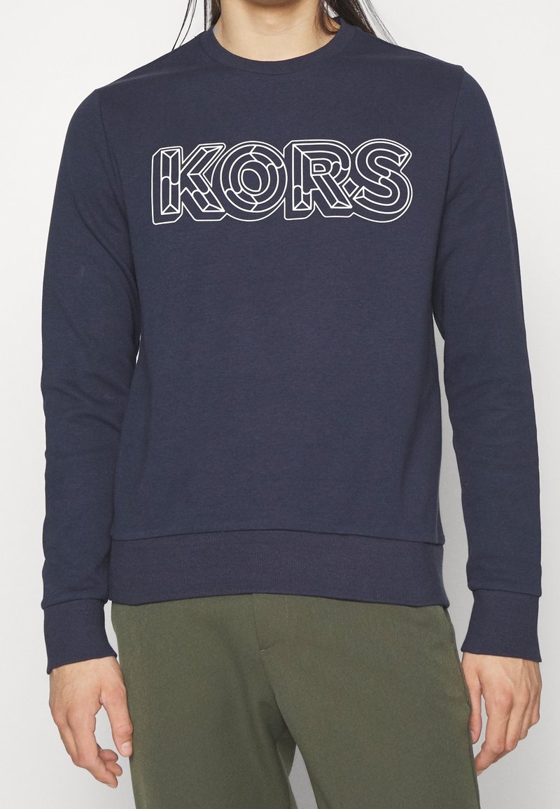 Granatowa bluza wykonana z miękkiej bawełny, z wyrazistym białym logo "KORS" w warstwowym wzorze, ściągaczami na mankietach i na dole.
