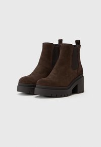 Bottines en suède marron avec des panneaux élastiques sur les côtés, bout rond et une semelle épaisse noire. Dispose d'une languette à l'arrière pour un enfilage facile.
