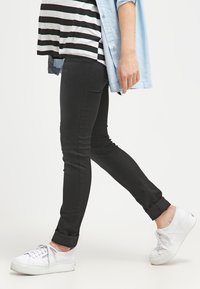 Jeans skinny pretos com um leve brilho, apresentando as bainhas dobradas. Combinados com uma camisa de botão azul claro sobre uma camisola riscada e ténis brancos.