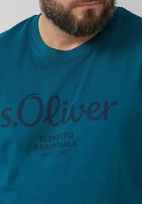 Turquoise katoenen t-shirt met een ronde hals, korte mouwen en grote bedrukte tekst: "s.Oliver ELEVATED ESSENTIALS SINCE 1969."