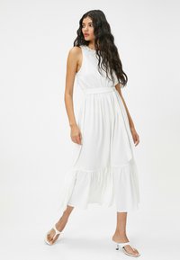 Robe blanche sans manches avec un corsage texturé, une taille cintrée et un ourlet à volants en couches. Portée avec des sandales à talons blanches.