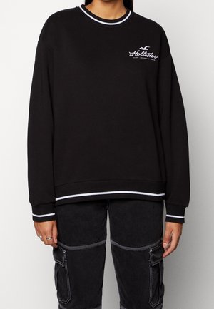Person iført en sort Hollister-sweatshirt med hvide stribede manchetter og kant, kombineret med sorte cargobukser med hvide syninger.