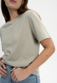 Hellgrünes Baumwoll-T-Shirt mit kurzen Ärmeln, Rundhalsausschnitt und lässiger Passform. Getragen mit hochgeschnittenen Jeansshorts, die ein dezentes Armtattoo zeigen.