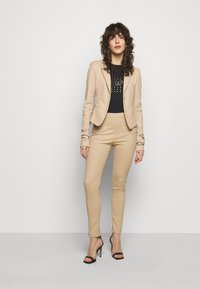 Blazer beige avec une coupe cintrée et une fermeture à un bouton, associé à un pantalon beige slim. Chemise noire avec des détails métalliques. Talons aiguilles noirs à lanières.