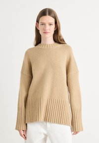 Beige oversized gebreide trui met een geribde zoom en manchetten, voorzien van een ronde halslijn en een gestructureerde stof. Gestyled met witte broeken.