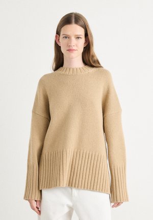 Beige oversized gebreide trui met een geribde zoom en manchetten, voorzien van een ronde halslijn en een gestructureerde stof. Gestyled met witte broeken.