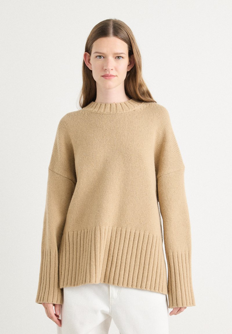 Beige oversized gebreide trui met een geribde zoom en manchetten, voorzien van een ronde halslijn en een gestructureerde stof. Gestyled met witte broeken.