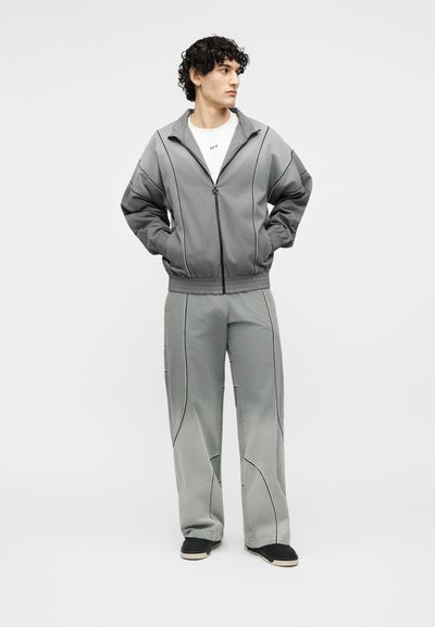 Νεαρός άνδρας με σγουρά μαλλιά στέκεται με τα χέρια στις τσέπες, φορώντας ταιριαστό γκρι ζακέτα με φερμουάρ και παντελόνι με φαρδιά μπατζάκια, και μαύρα παπούτσια.