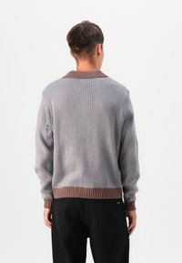 Knitted sweater in gray with a textured pattern, featuring a brown ribbed collar and hem, shown from the back with black pants.