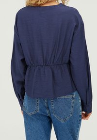 Blusa peplum blu navy con maniche lunghe, tessuto testurizzato e vita elastica. Abbinata a jeans blu a vita alta con tasche posteriori.