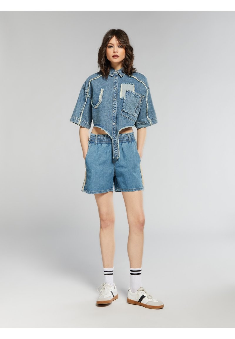 Camicia di jeans corta con maniche corte e tasca frontale, abbinata a pantaloni corti in denim con vita elastica. Entrambi sono di un blu chiaro con bordi sfilacciati.