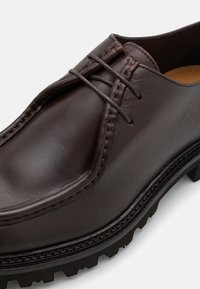 Chaussure en cuir marron avec un motif cousu, dotée d'un bout arrondi et d'une semelle en caoutchouc épaisse noire avec des rainures. Les lacets sont bien visibles.