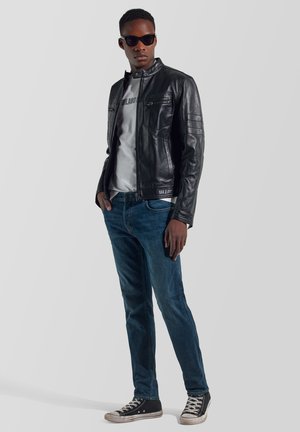 Joven con chaqueta de cuero negra, camiseta gráfica blanca, vaqueros azules, zapatillas negras altas y gafas de sol oscuras, de pie frente a un fondo liso.