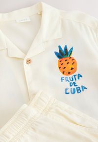 Cămașă de culoare crem cu o grafică ananas și text albastru "FRUTA DE CUBA" pe piept, asortată cu pantaloni crem.