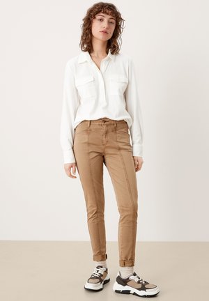 Beige enganliegende Hose mit seitlichen Nähten, kombiniert mit einem weißen Langarmhemd und gemusterten Sneakers. Neutraler Hintergrund.