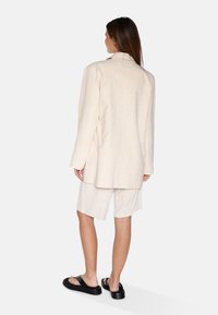 Világos bézs vászon blazer megfelelő rövidnadrággal, amely laza szabású. Fekete lapos szandálok egészítik ki a szettet. Minimalista design.