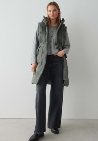 Gepolsterte ärmellose Weste in gedämpftem Grün, mit Kapuze, Reißverschluss und Taschen, über einem hellgrauen Hemdstrickpullover und einer dunklen Jeans getragen.