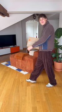 Καφέ καναπές από κορντόρι με πορτοκαλί λεπτομέρειες, ξύλινο πάτωμα, πράσινο φυτό σε γλάστρα και μια φιγούρα που κρατάει ένα χαρτόκουτο σε έναν φωτεινό, μοντέρνο χώρο.