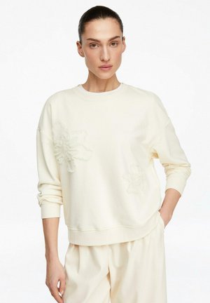 Donna che indossa una felpa color crema e pantaloni abbinati, con dettagli floreali in rilievo sulla felpa, in piedi davanti a uno sfondo bianco.