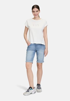 T-shirt en coton blanc à manches courtes avec des broderies ludiques, associé à un short en denim bleu clair et des baskets de sport grises.