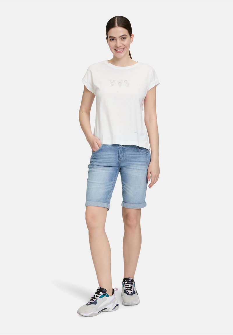 Witte katoenen t-shirt met korte mouwen en speelse borduursels, gecombineerd met lichtblauwe denim shorts en grijze sportieve sneakers.