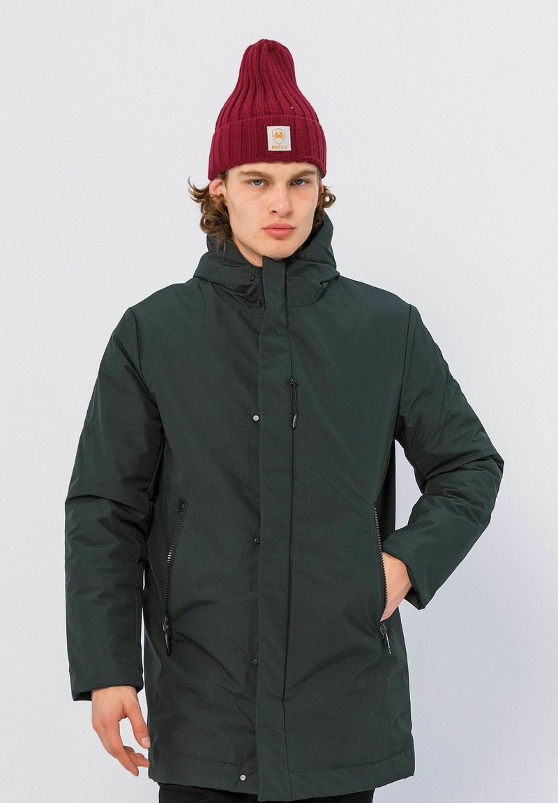 Donkergroene gewatteerde jas met een hoge kraag, ritszakken en een zachte textuur. Gekoppeld met een bordeauxrood gebreid mutsje.