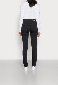 Jeans negros ajustados con cintura alta y detalles en los bolsillos traseros, combinados con una blusa blanca de manga larga y zapatillas blancas.