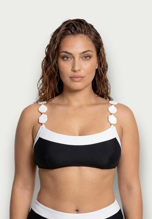 Mujer con cabello castaño ondulado lleva un top de bikini en blanco y negro con acentos decorativos de tirantes geométricos blancos, mirando hacia adelante sobre un fondo liso.