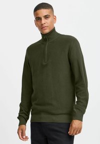 Olivgrüner Strickpullover mit hohem Kragen und Reißverschluss, versehen mit strukturierter Oberfläche sowie gerippten Bündchen und Saum. Sichtbare Details mit schuppigem Muster.