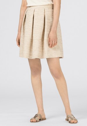 Frau trägt einen beigen, strukturierten, plissierten Rock, ein cremefarbenes Oberteil und goldene offene flache Sandalen, steht vor einem schlichten Hintergrund.