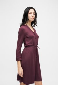 Γυναίκα που στέκεται με τα χέρια χαμηλά, φορώντας ένα μακριάς μανίκιας, μέχρι το γόνατο, μπουτόν φόρεμα σε χρώμα δαμάσκηνου με ζώνη στη μέση, σε απλό φωτεινό φόντο.