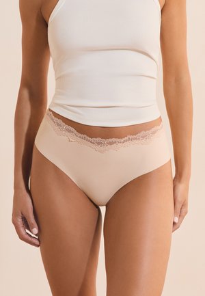 Mujer con ropa interior beige de corte alto con encaje y una camiseta de canalé blanca sin mangas sobre un fondo neutro.