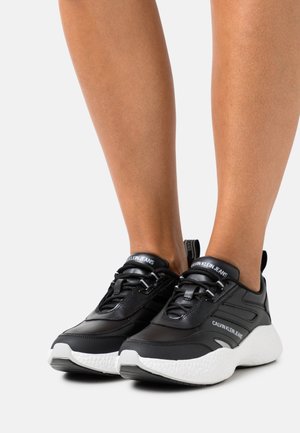 Chaussures de sport noires au design élégant, dotées d'une empeigne texturée, d'une semelle blanche et d'accents de marque sur la languette et le côté.