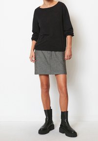 Schwarzes Langarmshirt mit rundem Ausschnitt, kombiniert mit einem strukturierten grauen Rock. Das Modell trägt klobige schwarze Stiefeletten.