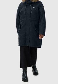 En person klädd i en marinblå parkas med pälsluva, svarta vida byxor och svarta läderstövlar som står mot en neutral bakgrund.