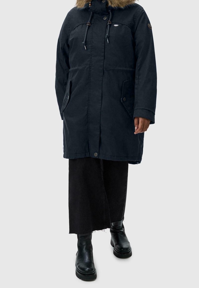 En person klädd i en marinblå parkas med pälsluva, svarta vida byxor och svarta läderstövlar som står mot en neutral bakgrund.